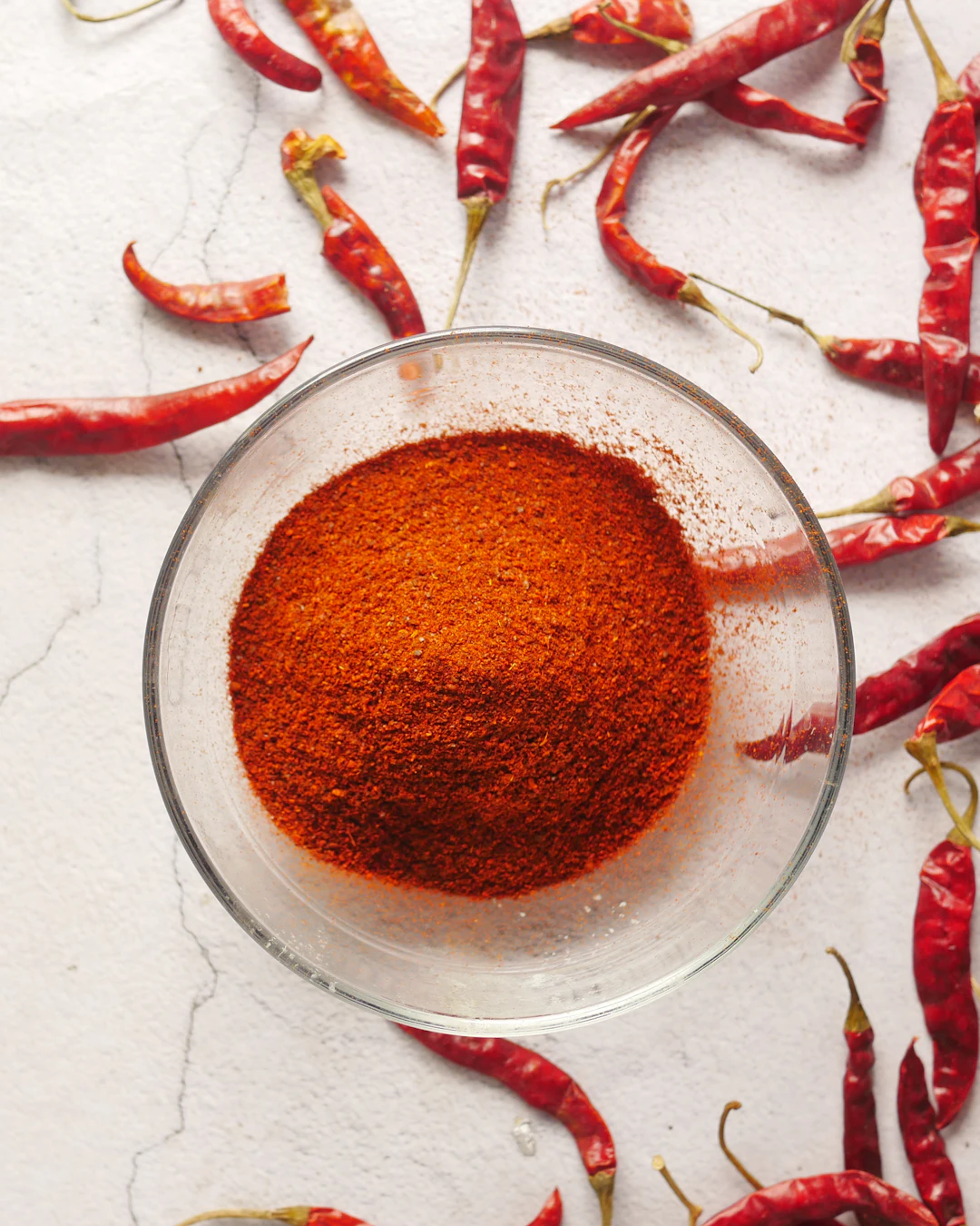 A glass bowl of red chilli powder surrounded by dried red chillies