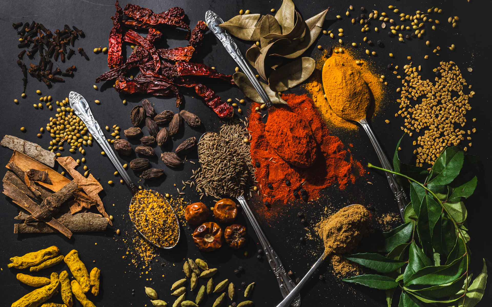 Whole and ground Indian spices — chilli powder, turmeric, dried red chillies and curry leaves — laid out on dark slate